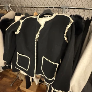 Svart jacka med vita detaljer - Säljer en unik svart jacka med vita broderade detaljer längs kanterna och fickorna. Jackan har en stilren design utan krage och långa ärmar. Perfekt för att ge en elegant touch till din outfit.