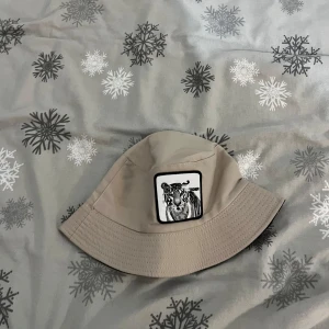 Beige bucket hat med tigerbroderi - Snygg beige bucket hat med ett coolt broderi av en tiger framtill. Perfekt för att ge din outfit en trendig touch. Hatten har en klassisk form och är lätt att matcha med olika stilar. Pris kan diskuteras! 