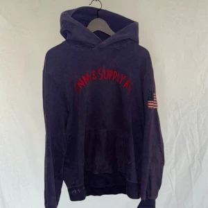 Mörkblå hoodie från Ralph Lauren - Säljer en snygg mörkblå hoodie från Ralph Lauren med röd text på bröstet och en amerikansk flagga på ärmen. Den har en klassisk känguruficka och ribbade muddar. Perfekt för en avslappnad stil. Marinblå men lite mera åt det lila hållet 