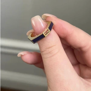 Guldfärgad ring med lila detalj från Fendi - Elegant guldfärgad ring från Fendi med en stilren lila detalj. Ringen har en modern design med en distinkt kantig form och Fendi-logotypen ingraverad. Perfekt för att ge en lyxig touch till din stil.