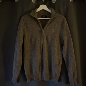 Grå ulltröja med dragkedja från GANT - Säljer en stilren grå tröja från GANT i extra fin lammull. Tröjan har en dragkedja framtill och broderad logga på bröstet. Perfekt för kyligare dagar med sin långa ärmar och mjuka material.