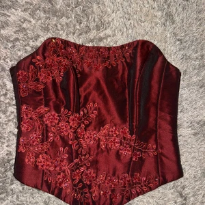 Vinröd korsett med blommig brodyr - Snygg vinröd korsett med vacker blommig brodyr och detaljer i siden. Använd en gång. Dragkedja bak. Storlek 36/38 alltså S/M pris kan diskuteras och skriv privat  ifall du vill ha fler bilder 