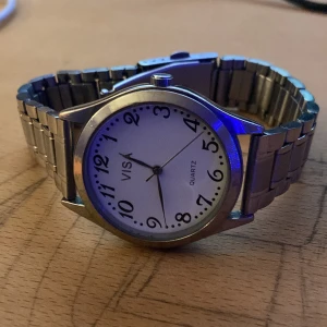 Vissa, klockan för 450 - Stilren herrklocka från Visa med en klassisk design. Klockan har en vit urtavla med svarta arabiska siffror och visare. Armbandet är i silverfärgat metall med en traditionell länkdesign. Perfekt för den som gillar en tidlös look.