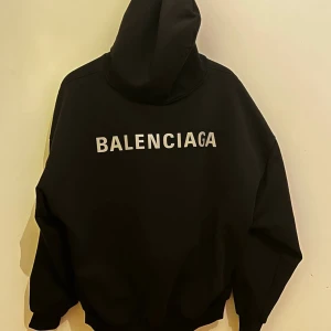 Svart hoodie från Balenciaga - Snygg svart hoodie från Balenciaga med logga i vitt på bröstet och ryggen. Den har en klassisk design med huva och en stor ficka framtill. Perfekt för en stilren och avslappnad look.