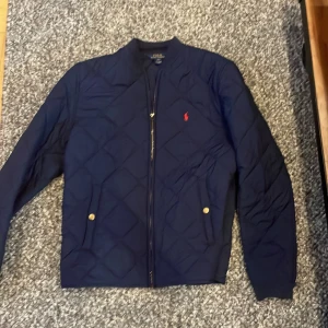 Mörkblå quiltad jacka från Ralph Lauren - Säljer en snygg mörkblå quiltad jacka från Ralph Lauren med dragkedja och två fickor med knappar. Den har en broderad röd logga på bröstet och långa ärmar. Perfekt för den kommande våren. Storleken är XL i barn storlek så skulle säga att den passar runt 170-180