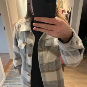 Rutig jacka från Lindex - Säljer en snygg rutig jacka från Lindex i beige, grå och brun. Jackan har långa ärmar och knappar framtill. Perfekt för höst och vår med sin mjuka och bekväma design. Två bröstfickor med lock ger en cool touch.