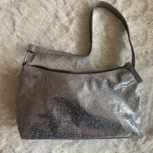 Glittrig silver axelväska från Monki - Snygg glittrig axelväska i silver från Monki. Väskan har en dragkedja upptill och en bekväm axelrem. Perfekt för att lägga till lite bling till din outfit. Den är kompakt men rymmer det viktigaste du behöver ha med dig.