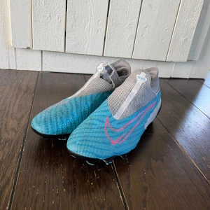 Ett par Nike Phantom GT fotbollsskor - Säljer ett par Nike Phantom GT fotbollsskor i blått och grått. Skorna har en modern design med snörningsfri ovandel och tydliga Nike-loggor. Perfekta för fotbollsplanen med sin strömlinjeformade passform. Skorna har ett hål på vristen (se bild) men inget som borde påverka spelet, den har även ett litet i vardera sula. Hör av er om ni har några frågor