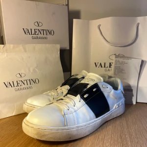 Valentino open sneakers - Säljer ett par riktigt snygga, vita Valentino Garavani sneakers med en svart detalj på sidan. Skorna har snörning och en klassisk design som passar perfekt för en stilren look. Allt förutom kvittot på bilderna medföljer vid köp. Jag köpte de här på Plick för 2999 men de passade ej. Osäker på äktigheten då de som sagt är köpta här på Plick, men jag tror att de är äkta!