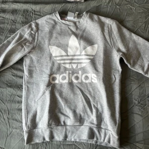 Grå sweatshirt från Adidas - Säljer en grå sweatshirt från Adidas med det klassiska trefoil-logotypen i vitt på framsidan. Tröjan har långa ärmar och en rund halsringning. Perfekt för en avslappnad stil.