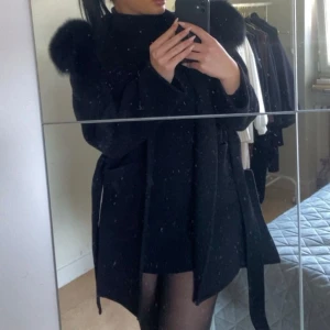 Svart kappa med pälskrage - Snygg svart kappa med stor pälskrage och knytband i midjan. Perfekt för kyliga dagar när du vill hålla stilen. Kappan har långa ärmar och en elegant design som passar till många olika outfits.