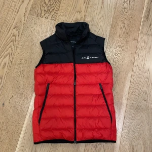 Röd och svart dunväst från Sail Racing - Snygg och sportig dunväst från Sail Racing i rött och svart. Västen har en dragkedja framtill och två praktiska fickor med dragkedja. Perfekt för kyliga dagar när du vill hålla stilen. 🖤❤️Säljer pga tröttnat, använd 3 gånger!