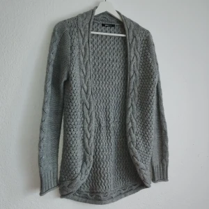 Grå stickad kofta från Gina Tricot - Mysig grå stickad kofta från Gina Tricot med flätstickade detaljer längs kanterna. Perfekt för kyliga dagar med sin långa design och bekväma passform. Ett måste i garderoben för en stilren look.