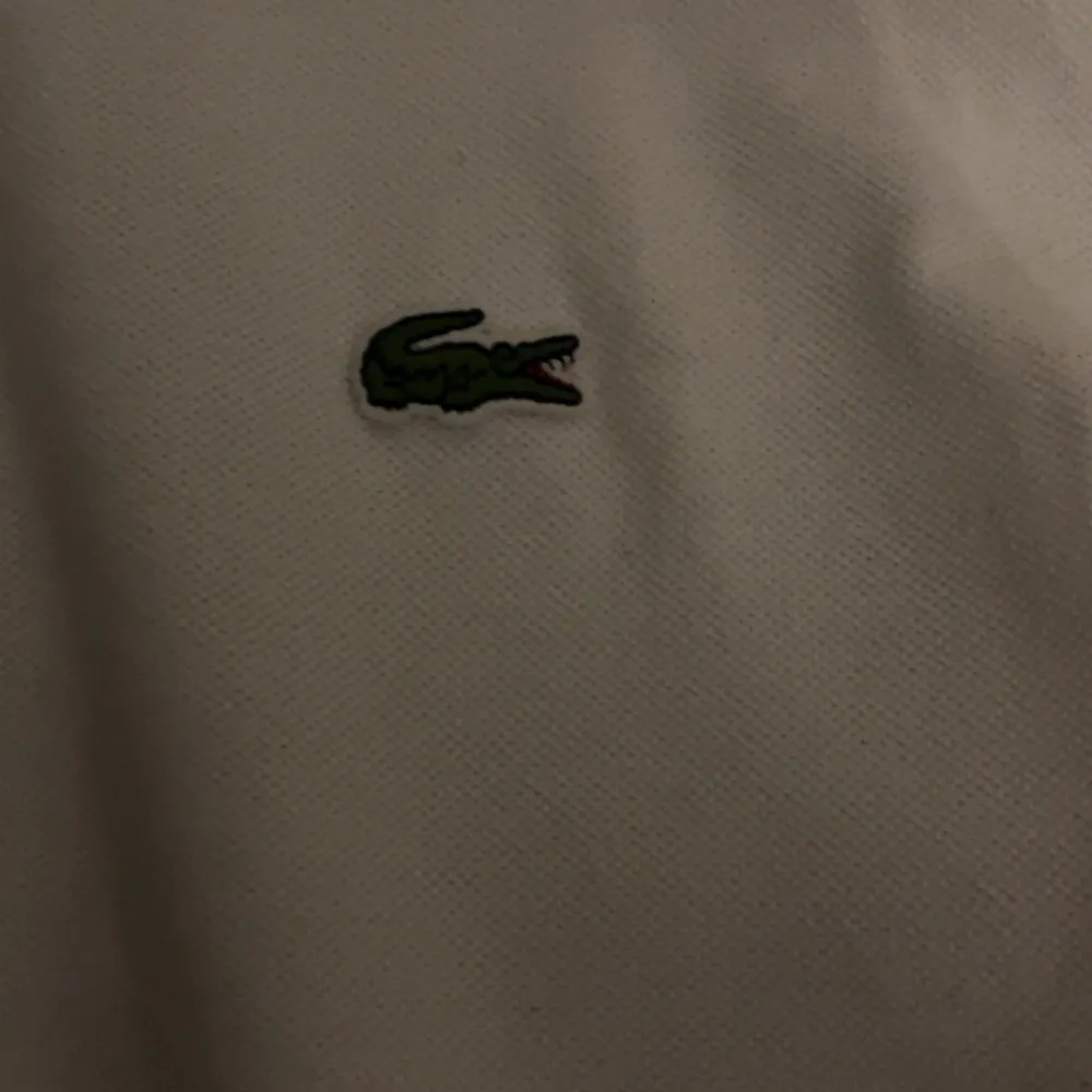 Säljer en klassisk vit pikétröja från Lacoste med den ikoniska gröna krokodilen på bröstet. Tröjan har korta ärmar och en traditionell krage med knappar. Perfekt för en stilren och avslappnad look.. Neuletakit & Villapaidat.