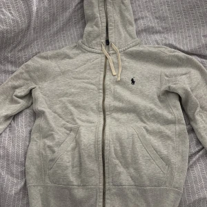 Grå hoodie från Ralph Lauren - Säljer en stilren grå hoodie från Ralph Lauren med dragkedja och huva. Tröjan har en liten broderad logotyp på bröstet och praktiska fickor framtill. Perfekt för en avslappnad look.