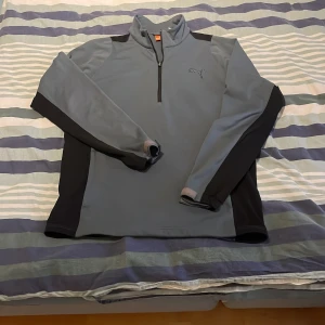 Grå tröja med dragkedja från Puma - Snygg grå tröja från Puma med halv dragkedja och svarta detaljer på ärmarna. Perfekt för träning eller en avslappnad dag. Tröjan har en diskret logga på bröstet och en bekväm passform. Vindjacka från puma. Pris går alltid att förhandlas!