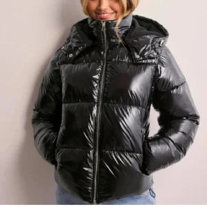 Nelly svart glansig jacka  - Snygg svart glansig pufferjacka med dragkedja och huva. Perfekt för kyliga dagar med sin vadderade design som håller dig varm och bekväm. Jackan har en modern look och passar bra till olika outfits. använd fåtal gånger därmed inga tecken på användning!😇🫶