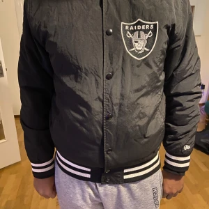 Svart bomberjacka med Raiders-logga - Snygg svart bomberjacka från New Era med Raiders-logga på bröstet och NFL-märke på ärmen. Jackan har knappar framtill och ribbade muddar med vita ränder vid ärmslut och nederkant. Perfekt för sportfantasten!