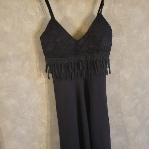 Svart klänning med spetsdetaljer - Elegant svart klänning med spetsdetaljer på överdelen och smala axelband. Klänningen har en v-ringad design och dekorativa fransar under bysten. Perfekt för en stilren look.