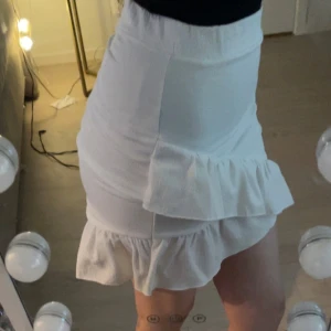 Vit kjol med volangdetaljer - Säljer en superfin vit kjol med volanger. Den har en omlott-design och är perfekt för sommaren. Kjolen är kort och har en elastisk midja för en bekväm passform. Perfekt för både vardag och fest! Säljer för att den inte kommer till användning! Den har använts på en skolavslutning men är relativt ny! 