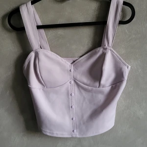 Ljuslila korsettopp med knappar - Snygg ljuslila korsettopp från Cropp med breda axelband och dekorativa knappar framtill. Perfekt för en stilren look.