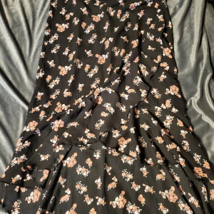 Svart blommig kjol - Snygg svart kjol med ett blommigt mönster i rosa och vitt. Kjolen har en volangdetalj som ger den en extra touch. Perfekt för en stilfull look.