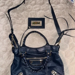 Mörkblå balenciaga city väska med detaljer - Snygg mörkblå axelväska i skinn med flätade handtag och guldfärgade metalldetaljer. Väskan har en praktisk dragkedja och justerbar axelrem. Perfekt för att bära dina dagliga nödvändigheter med stil. Balenciaga City