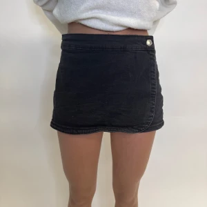 Svart jeanskjol - Trendig svart jeanskjol med omlott-design och knappdetalj i midjan. Kjolen har en kort längd och två bakfickor som ger en cool look. Perfekt för att matcha med en enkel topp eller tröja. Framifrån ser det ut och vara en kjol men är egentligen shorts. Som referens är modellen 164cm lång och brukar vanligtvis bära storleken 34.