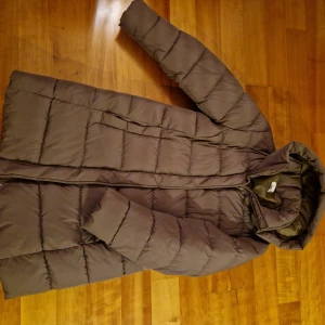 Brun/Grön dunjacka från H&M - Säljer en lång dunjacka från H&M med huva. Jackan har en dragkedja framtill och är quiltad för extra värme. Perfekt för kyliga dagar. Rök och djurfri. Ej använd så mycket.