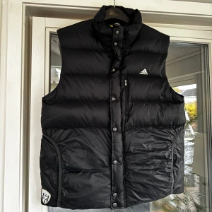 Svart dunväst från Adidas - Snygg svart dunväst från Adidas med dragkedja och knappar. Perfekt för kyligare dagar med sin quiltade design. Västen har en hög krage och praktiska fickor framtill. Kika och se om du ser något mer i min profil som du gillar så skickar jag med det till dig också!😊 Fler kläder kommer! 