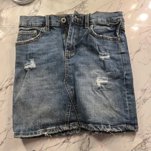 Jeanskjol med slitningar - Snygg jeanskjol i blå denim med slitna detaljer och rå kant. Kjolen har en klassisk femficksdesign och stängs med knapp och dragkedja framtill. Perfekt för en avslappnad stil.