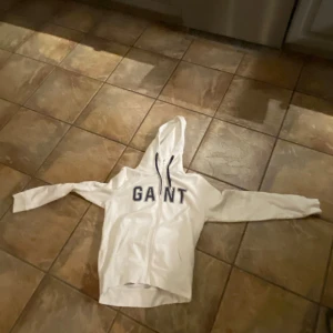 Vit hoodie från GANT - Säljer en vit hoodie från GANT med dragkedja och stor logga på framsidan. Perfekt för en avslappnad look. Den har långa ärmar och en bekväm huva. Några delar av tröjan är det grön/blå prickar 