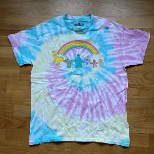 Tai-dye t-shirt med Sesame Street - Cool t-shirt med tie-dye mönster i blått, rosa och gult. Framsidan har en regnbåge och karaktärer från Sesame Street. Baksidan har texten 'L IS FOR LOVE'
