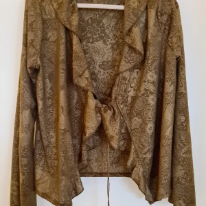 Brun blus från Zara - Snygg brun blus från Zara med blommigt mönster och knytning framtill. Blusen har långa ärmar och volangdetaljer längs kragen. Perfekt för en stilfull look till festen! Nypris 499.