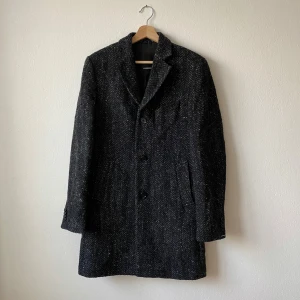 Svartvit Wolger rock från J.Lindeberg - Elegant svart kappa från J.Lindeberg i ullblandning. Klassisk design med knappar framtill och en stilren krage. Perfekt för kyligare dagar med sin långa passform och fodrade insida. Modell: Wolger 77 rock Wool - Artnr:G097