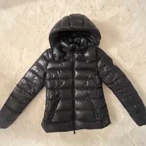 Svart dunjacka från Moncler - Snygg svart dunjacka från Moncler med glansig finish. Jackan har en dragkedja framtill och en praktisk huva. Perfekt för kyliga dagar. Tveka inte att höra av er om ni har frågor!!💓💓 