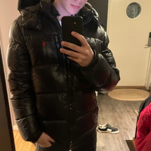 Svart dunjacka från Ralph lauren - Snygg svart dunjacka med huva och dragkedja framtill. Jackan har en broderad logotyp på bröstet och praktiska fickor med dragkedja. Perfekt för kyliga dagar. Bra skick bara väldigt liten defekt syns på sista bild, självklart äkta i storlek S!!