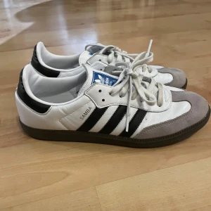 Adidas Samba sneakers - Klassiska Adidas Samba sneakers i vitt och svart med grå detaljer. Har använt skorna i 1 säsong, därmed har skorna inga skador eller defekter. Köpte dom förra året men har vuxit ur dom, så därför väljer jag att sälja dom.