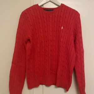 Röd kabelstickad tröja från Ralph Lauren Sport - Säljer en snygg röd kabelstickad tröja från Ralph Lauren Sport. Tröjan har en klassisk design med rund hals och det ikoniska broderade logotypen på bröstet. Perfekt för kyligare dagar.