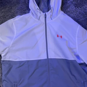 Vit och grå vindjacka från Under Armour - Jackan är L men passar även M, Snygg vindjacka från Under Armour i vitt och grått med huva och dragkedja. Perfekt för blåsiga dagar. Den har en röd logotyp på bröstet och en praktisk design.
