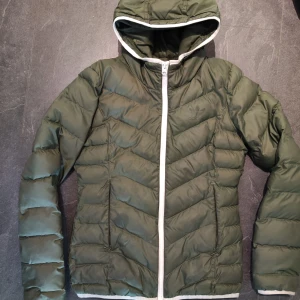 Grön dunjacka från Adidas, strl XS - Säljer en snygg grön dunjacka från Adidas med vit dragkedja och huva. Jackan har en quiltad design och är perfekt för kyligare dagar. Den är långärmad och har en sportig look. Har ett litet märke på ärmen men går att dölja lätt för den fixiga.