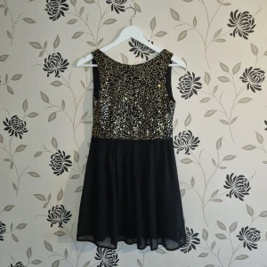 Svart klänning med guldpaljetter Strl: 36 - Snygg svart kortklänning från Elise Ryan med glittriga guldpaljetter på överdelen. Klänningen är ärmlös och har en öppen rygg som ger en elegant touch. Perfekt för festliga tillfällen.