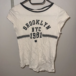 Vit t-shirt med Brooklyn-tryck - Snygg vit t-shirt med svart text 'Brooklyn NYC 1981' och ett litet hjärta. Den har korta ärmar och en rund halsringning med svart kant. 