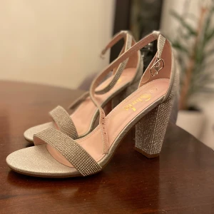 Glittriga sandaler med klack - Snygga sandaler med hög klack i glittrigt material. De har en elegant remdesign och en justerbar spänne runt ankeln. Perfekta för att ge en touch av glamour till din outfit. 