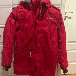 Röd parkajacka från Didriksons - Snygg röd parkajacka från Didriksons med pälsfodrad huva och praktiska fickor. Jackan har dragkedja och knappar framtill för extra skydd mot kyla. Längst ner på dragskedjan har vi behövt sy om då den gått sönder men ingen som syns när man har på sig den, sedan är det också en knapp som är borta men går att fixa om man är händig, annars inga mer defekter.