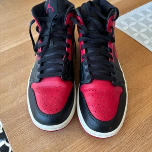 Nike Air Jordans 1 mid - Snygga Nike Air Jordans i klassisk svart och röd färgkombination. Skorna har en mid modell med snörning och den ikoniska Air Jordan-loggan på sidan. Perfekta för dig som vill ha en stilren och sportig look.