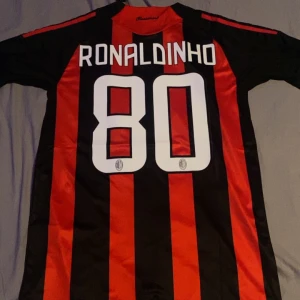 AC Milan fotbollströja med Ronaldinho 80 - Snygg AC Milan fotbollströja från Adidas med Ronaldinho och nummer 80 på ryggen. Tröjan är svart och röd med korta ärmar och har Bwin som sponsorlogga på framsidan. Perfekt för fans av Ronaldinho och AC Milan!