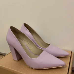 Ljuslila pumps med klack från Nelly - Snygga ljuslila pumps med spetsig tå och blockklack. Perfekta för att ge en färgklick till din outfit. Tillverkade i syntetmaterial.