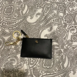 Svart plånbok från Ralph Lauren - Snygg och praktisk svart plånbok från Ralph Lauren med gulddetaljer. Den har flera kortfack och en dragkedja för säker förvaring. Perfekt för att hålla ordning på dina kort och kontanter. Nypris på denna är 1095:-