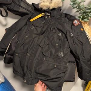 Svart parkajacka med pälsdetalj - Snygg svart parkajacka från Parajumpers med pälsdetalj på huvan. Jackan har flera praktiska fickor med dragkedjor och knappar, samt en broderad logga på ärmen. Perfekt för kalla dagar. Säljs pga att jag valt att köpa en ny jacka Storlek L vid frågor hör av er.